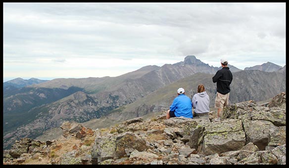 Hikers on top of Hallett Peak looking towards Longs Peak under light grey skies in Rocky Mountain National Park
