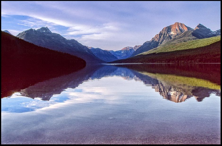 The surrounding mountain peaks reflect in Bowman Lake in Glacier National Park, Montana and Psalm 93:1 Bible verse robed in majesty