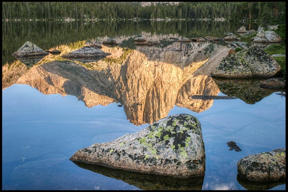 Mountains and a blue sky reflect next to a rock in Middle Cathedral Lake, Wind Rivers Wilderness, Wyoming and Bible verse Psalm 103:21-22 Works of His