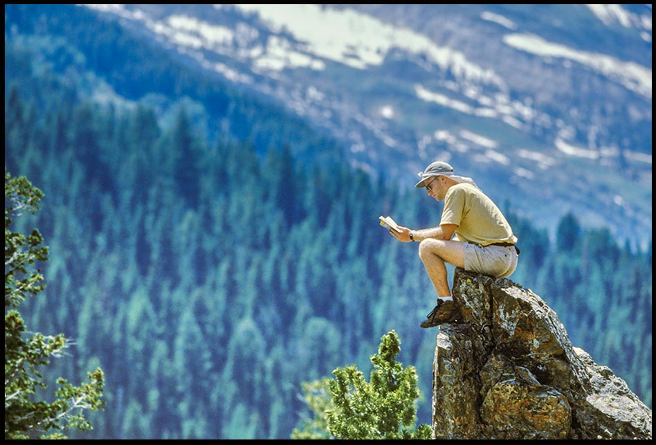 A hiker on a sitting the peak of large boulder, White River National Forest, Colorado. Bible Verse of the Day: Psalm 19:4