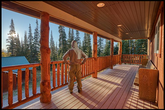 A man standing on a cabin deck in a pine forest watching the sunrise. Draw near to God at the table in the wilderness camp in Wyoming