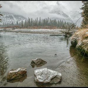 Rocks in slow moving river surrounded cloud covered Canadian Rockies in winter, Alberta, Canada. Psalm 46:4-5 Bible verse God in our Midst